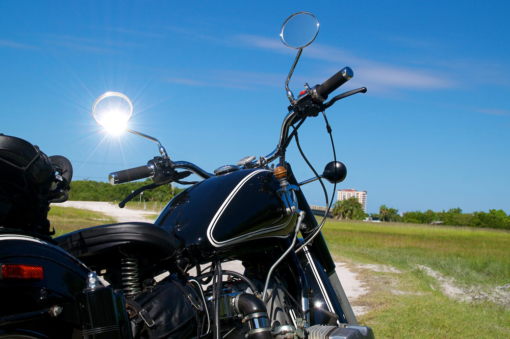 A black vintage motorcycle on a dirt road under a sunny sky with glare on mirror.